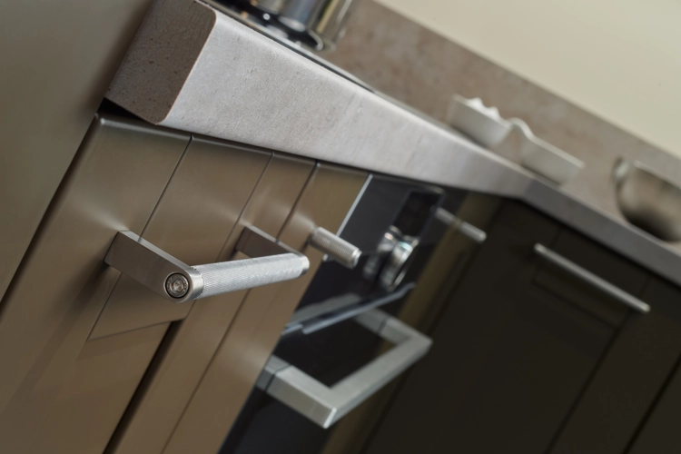 Detail moderní kuchyně s kovovými úchyty na matných skříňkách v tmavém odstínu. Kamenná pracovní deska dodává interiéru elegantní vzhled. Vestavná trouba s nerezovým madlem ladí s industriálním stylem kuchyně.
