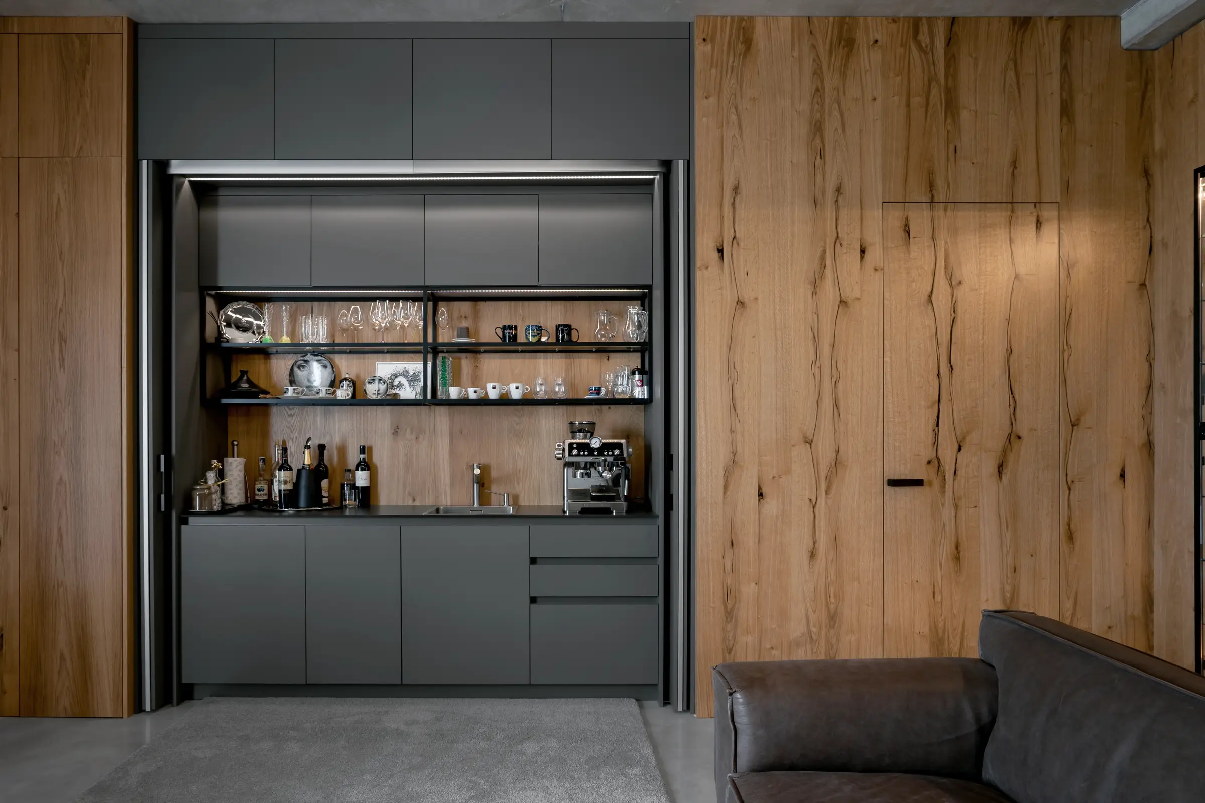 Moderní kuchyně s tmavě šedými skřínkami a dřevěnou stěnou v kombinaci s otevřenými policemi plnými sklenic, hrnků a lahví. Na pulce stojí kávovar a další kuchynské vybavení, v popředí je část obývacího pokoje s pohovkou a šedým kobercom.