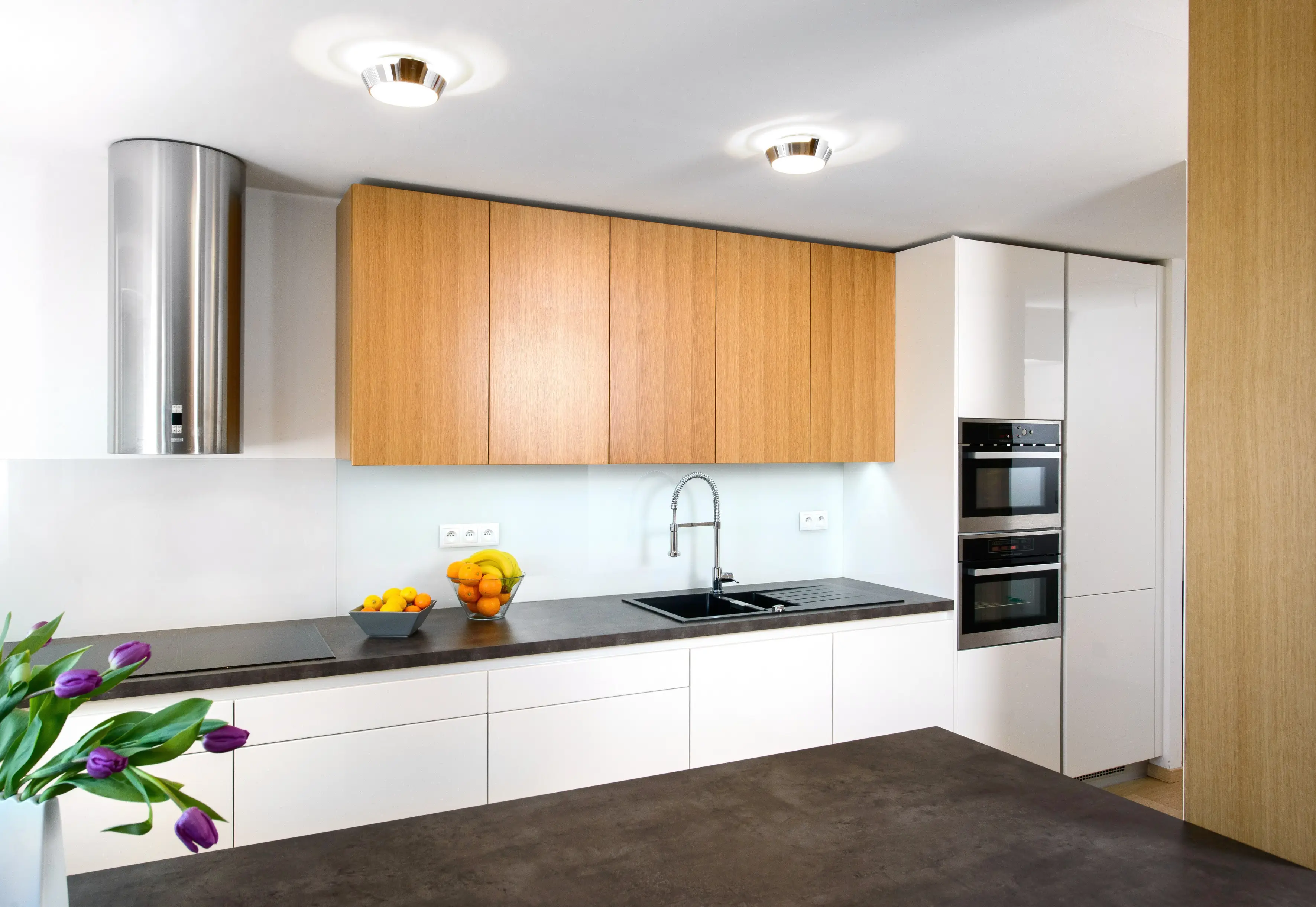 Moderní kuchyň s bílými spodními skříňkami, horními skříňkami v dekoru světlého dřeva a tmavou pracovní deskou. Kuchyňská linka je doplněna vestavnou troubou, černým dřezem s vysokou baterií a minimalistickým osvětlením.